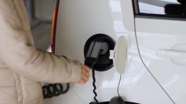Kadın bir arabanın önünü açıyor ve elektrik kablosunu kamu şarj noktasında elektrikli araçtan çıkarıyor..