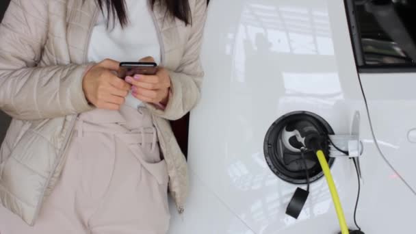 Женщина со смартфоном возле подзарядки электромобиля и проверки состояния аккумулятора. Автомобиль зарядки на общественной зарядной станции на открытом воздухе. Концепция совместного использования автомобилей — стоковое видео