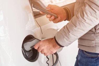 Elektrikli bir arabayı şarj eden ve gün ışığında bu akıllı telefonun aküsünü kontrol eden bir adam. Araba paylaşımı veya elektrikli araç kiralama. 