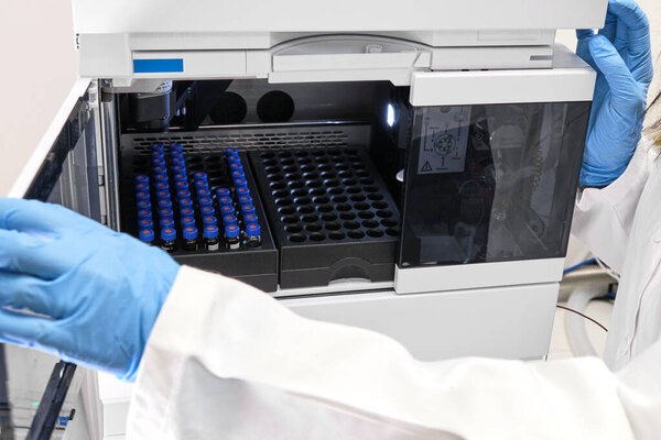 Scientist in a white lab coat putting vial with a sample into autosampler of HPLC system. High performance liquid chromatography at chemical laboratory. Developing of pharmaceuticals or vaccine