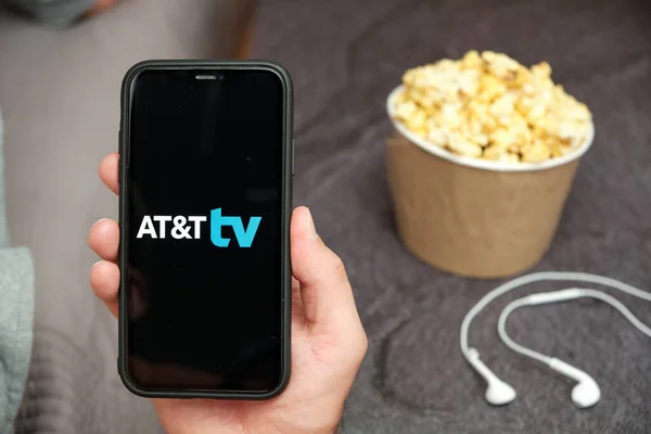 Close up mans hand holding a mobile phone with AT and T TV logo with Apple earphones and popcorn box next by him, Free TV concept, August 2020, San Francisco, USA — Stockfoto