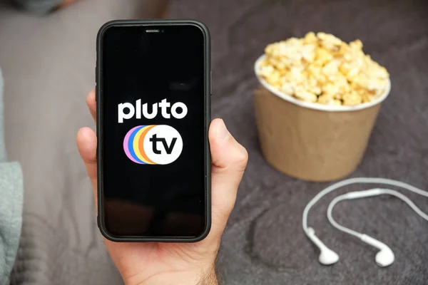 Yanında Apple kulaklıklar ve patlamış mısır kutusu olan Plüton TV logosu olan bir cep telefonu taşıyan bir adamın elini kapatın. Ücretsiz TV konsepti, Ağustos 2020, San Francisco, ABD