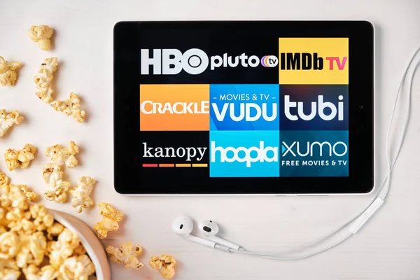 Movies Applikationslogo auf dem Bildschirm des Tablets auf dem weißen Tisch liegend und mit Popcorn bestreut. Apple-Kopfhörer in der Nähe des Tablets mit HBO, pluto, IMDb TV, Crackle, Vudu, tubi, kanopy — Stockfoto
