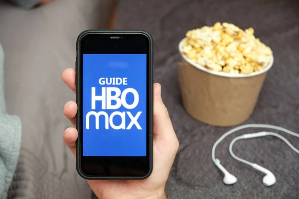 Close up mans hand holding a mobile phone with Guide HBO max logo mit Apple-Kopfhörern und Popcorn-Box neben ihm, August 2020, San Francisco, USA — Stockfoto