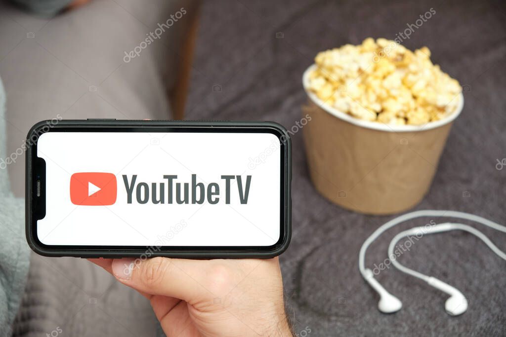 Close up mans hand holding a mobile phone with YouTube TV logo with Apple earphones and popcorn box next to him, Free TV concept, August 2020, San Francisco, USA.