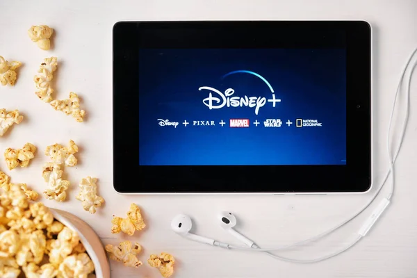 Logotipo de Disney plus en la pantalla de la tableta colocada en la mesa blanca y espolvoreado palomitas de maíz en ella. Auriculares Apple cerca de la tableta que muestra una aplicación de Disney, agosto 2020, San Francisco, EE.UU. —  Fotos de Stock