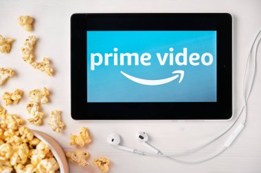Tabletin ekranındaki Amazon ana video logosu beyaz masaya serpilmiş ve üzerine patlamış mısır serpiştirilmiş. Tabletin yanındaki Apple kulaklıklar, Ağustos 2020, San Francisco, ABD