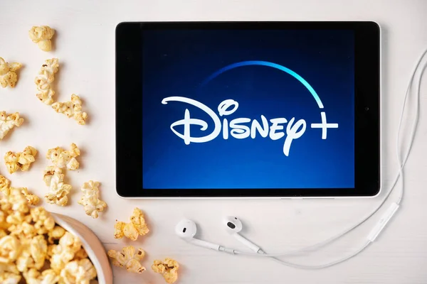 Logotipo de Disney plus en la pantalla de la tableta colocada en la mesa blanca y espolvoreado palomitas de maíz en ella. Auriculares Apple cerca de la tableta, agosto 2020, San Francisco, EE.UU. —  Fotos de Stock