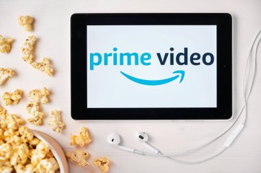 Tabletin ekranındaki Amazon ana video logosu beyaz masaya serpilmiş ve üzerine patlamış mısır serpiştirilmiş. Tabletin yanındaki Apple kulaklıklar, Ağustos 2020, San Francisco, ABD