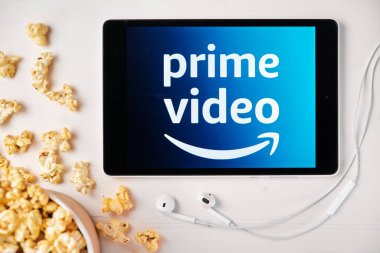 Tabletin ekranındaki Amazon ana video logosu beyaz masaya serpilmiş ve üzerine patlamış mısır serpiştirilmiş. Tabletin yanındaki Apple kulaklıklar, Ağustos 2020, San Francisco, ABD