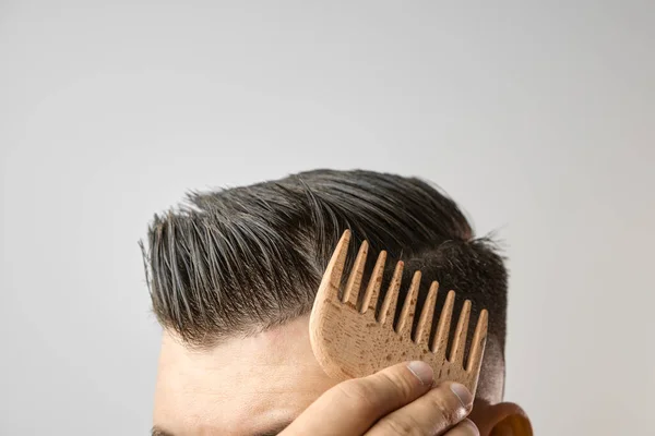 Mann bürstet sich die Haare mit einem Holzkamm und zeigt, wie man sich zu Hause um die Haare kümmert. Moderner und stilvoller Haarschnitt nach dem Friseur. — Stockfoto