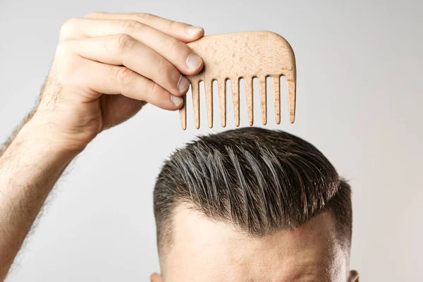 Mann bürstet sich die Haare mit einem Holzkamm und zeigt, wie man sich zu Hause um die Haare kümmert. Moderner und stilvoller Haarschnitt nach dem Friseur. — Stockfoto