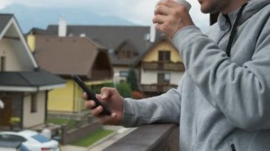 Akıllı telefonlu bir adam ve balkonda beyaz bir kupa ile harika bir dağ manzarası var..