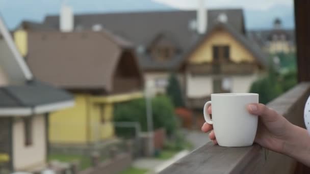 Cambiando el foco de la copa a las montañas. Acercamiento mujer sosteniendo una taza blanca en el balcón con vista a las casas modernas o pensiones y altas montañas. — Vídeos de Stock