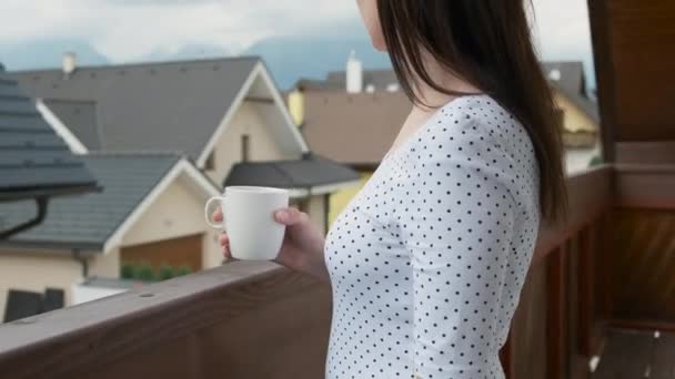 Acérquese a la joven bebiendo de una taza blanca en el balcón con una vista increíble de las montañas y casas nuevas. Mañana perfecta en el campo. — Vídeo de stock