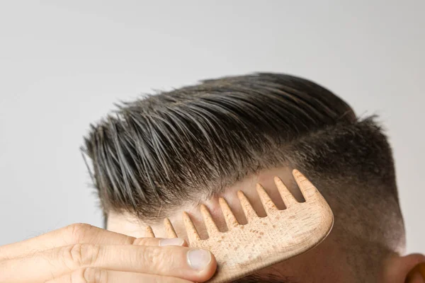 Mężczyzna z bliska szczotkujący włosy drewnianym grzebieniem na szarym tle. Leczenie włosów utraconych i łupieżu koncepcji. Produkty kosmetyczne dla mężczyzn. Reklama fryzjera. — Zdjęcie stockowe