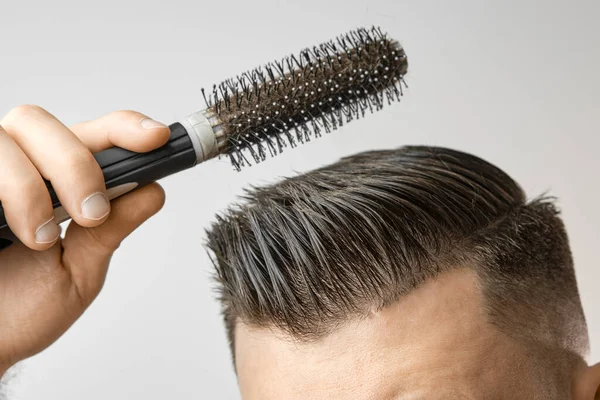 Der Mann benutzt eine runde Bürste, um seine Haare zu stylen. Haarpflege zu Hause nach dem Friseur. Kämmen der kurzen braunen Haare auf dem weißen Hintergrund — Stockfoto