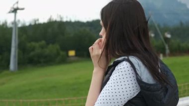 Sıcak bir yelek giymiş, konuşan akıllı telefonlu bir kadın dağların arka planında duruyor ve kayak asansörleri ya da fünikülerle hareket ediyor. Dağlarda bulutlu ve rüzgarlı bir hava var. Yürüyüşe çıkan bir turist. 