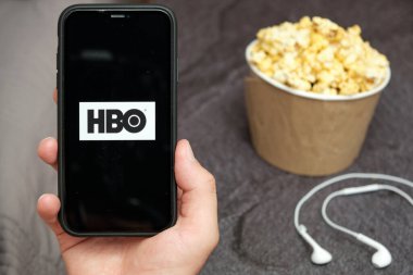 Yanında HBO logosu, Apple kulaklıkları ve patlamış mısır kutusu olan bir cep telefonu taşıyan bir adamın elini kapat, Ağustos 2020, San Francisco, ABD