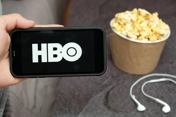 Close up mans hand holding a mobile phone with HBO logo with Apple Earphones and popcorn box next him, August 2020, San Francisco, USA — Stockfoto