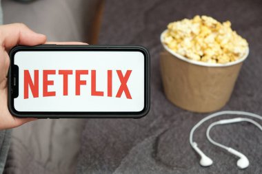 Netflix logosu, Apple kulaklıkları ve yanında patlamış mısır kutusu olan bir cep telefonu ile yakın çekim adamı, Ağustos 2020, San Francisco, ABD