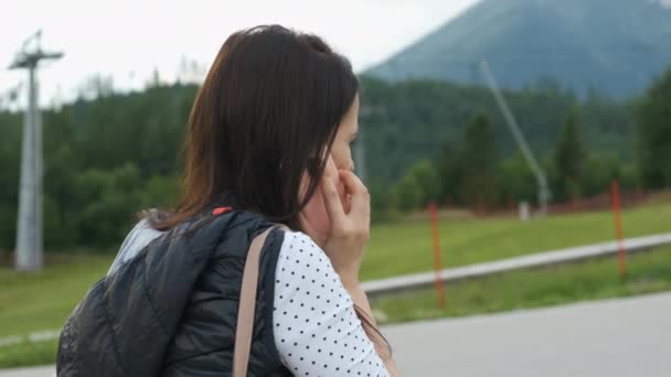 Kvinna med en ryggsäck på axeln i profil talar på smartphone med bergen på bakgrunden och flytta skidliftar. Turisten kommer att vandra — Stockvideo