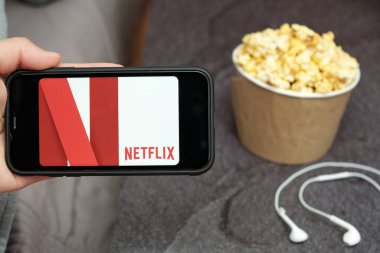 Netflix logosu, Apple kulaklıkları ve yanında patlamış mısır kutusu olan bir cep telefonu ile yakın çekim adamı, Ağustos 2020, San Francisco, ABD