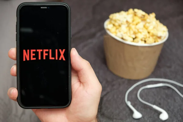 In Großaufnahme hält seine Hand ein Mobiltelefon mit Netflix-Logo, neben ihm Apple-Kopfhörer und Popcorn-Box, August 2020, San Francisco, USA — Stockfoto