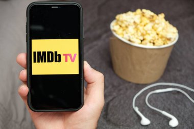 Yanında Apple kulaklıklar ve patlamış mısır kutusu olan IMDb TV logosu olan bir cep telefonu tutan adamları kapatın, Ağustos 2020, San Francisco, ABD
