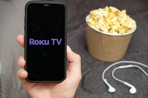 In Großaufnahme hält seine Hand ein Handy mit Roku-TV-Logo, daneben Apple-Kopfhörer und Popcorn-Box, August 2020, San Francisco, USA — Stockfoto