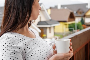Puantiyeli tişörtlü genç bir kadın balkonda beyaz bir fincan çay ya da kahve ile dikiliyor. Kırsal kesimde sabah. Rahatlama kavramı. 