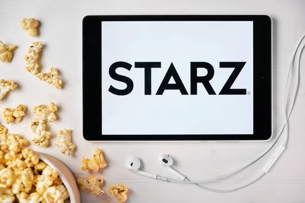 Logo de Starz en la pantalla de la tableta colocada en la mesa blanca con palomitas de maíz dispersas y auriculares Apple. Pasando tiempo libre en casa o anuncio de noticias, septiembre 2020, San Francisco, EE.UU.. —  Fotos de Stock