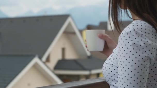 Close-up jonge vrouw drinken uit een witte beker op het balkon met een prachtig uitzicht op de bergen en nieuwe huizen. Perfecte ochtend op het platteland. — Stockvideo