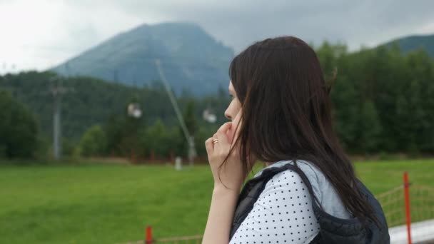 Kvinna i en varm väst talande smartphone som står på bakgrunden av berg och flytta skidliftar eller enhörningar. Molnigt och blåsigt väder i fjällen. Turist på vandring. — Stockvideo