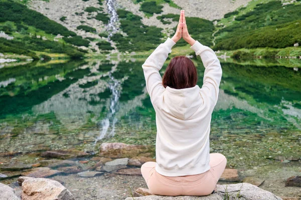 Young woman in a white hoodie practicing yoga on a lake shore with transparent water with an amazing view on waterfall. Meditation and recreation in nature between mountains.