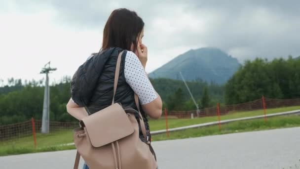 Vrouw met een rugzak op de schouder in profiel praten op smartphone met de bergen op de achtergrond en bewegende skiliften. Toerist gaat wandelen — Stockvideo