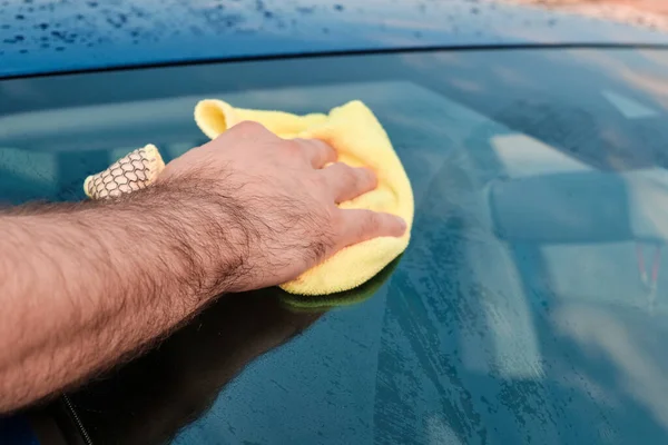 Close up mans hand polishing or cleaning a front windshield of a blue automobile with a yellow microfiber rag . Manual car washing.