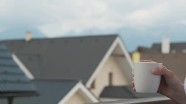 Zblízka mladá žena pije z bílého poháru na balkóně s úžasným výhledem na hory a nové domy. Perfektní ráno na venkově. — Stock video