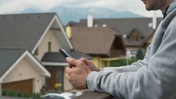 Ο άνθρωπος που χρησιμοποιεί το smartphone του στο μπαλκόνι με φόντο τα νέα σπίτια και τα βουνά. — Αρχείο Βίντεο