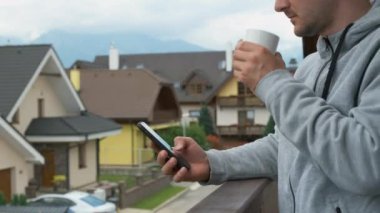 Akıllı telefonlu bir adam ve balkonda beyaz bir kupa ile harika bir dağ manzarası var..