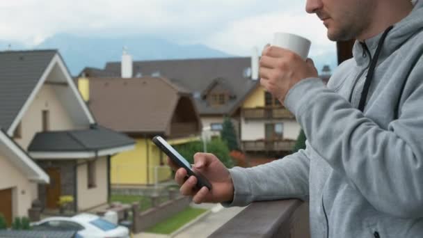 Uomo in piedi con uno smartphone e una tazza bianca sul balcone con una splendida vista sulle montagne. — Video Stock