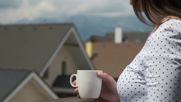 Nahaufnahme junge Frau trinkt aus einer weißen Tasse auf dem Balkon mit herrlichem Blick auf die Berge und neue Häuser. Perfekter Morgen auf dem Land. — Stockvideo