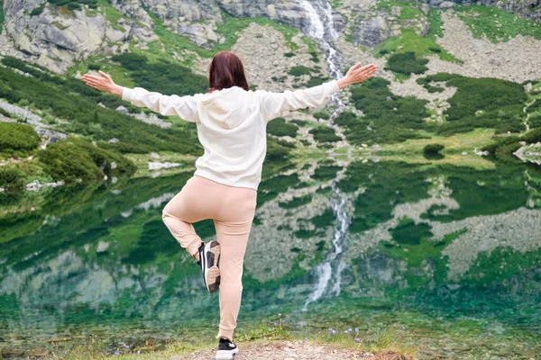 Женщина практикует йогу на берегу озера с удивительным видом на водопад. Расслабление и медитация о природе. — стоковое фото