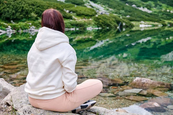 Молодая женщина сидит на берегу озера с прозрачной водой и наслаждается видом с копировальным пространством. Расслабление и медитация над концепцией природы. — стоковое фото
