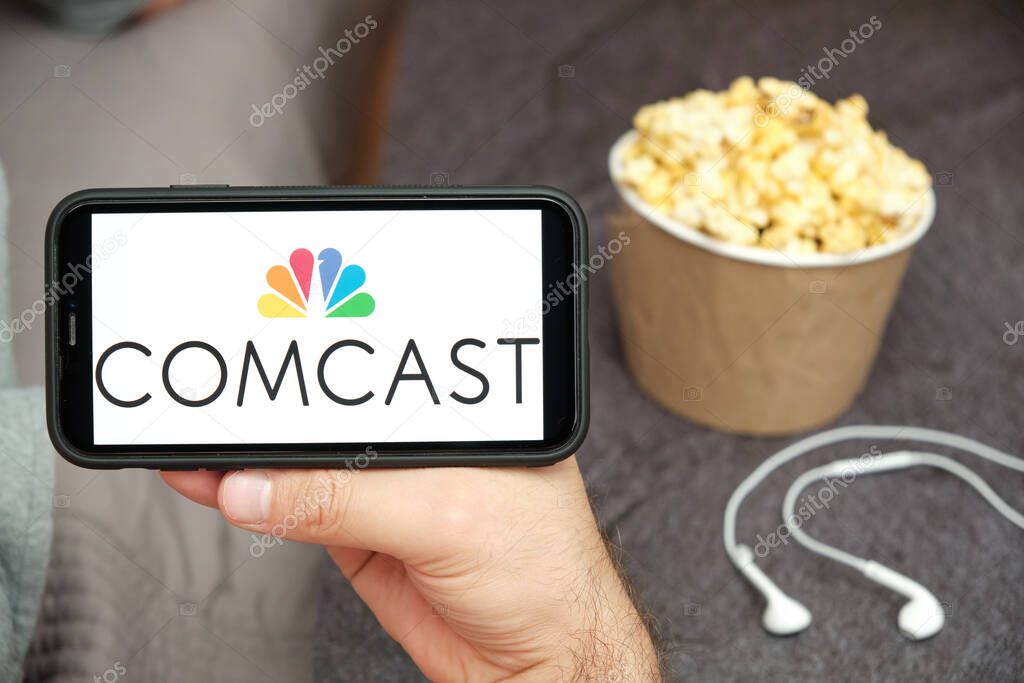 Comcast logo on the mobile phone screen with popcorn box and Apple earpods on the background. Leisure time at home concept. September 2020, San Francisco, USA.