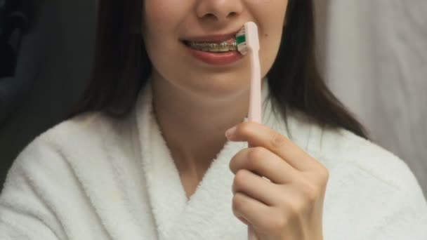 La filmación como correctamente limpiar los dientes con los aparatos ortopédicos por la mujer en el albornoz blanco en el reflejo del espejo — Vídeos de Stock