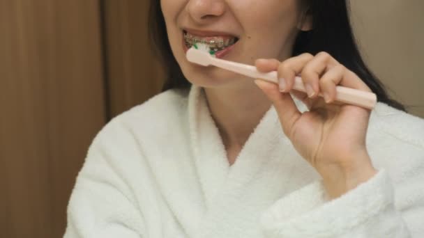 La filmación como correctamente limpiar los dientes con los aparatos ortopédicos por la mujer en el albornoz blanco en el reflejo del espejo. Higiene dental. — Vídeos de Stock