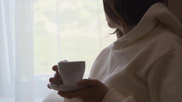 Młoda kobieta w białej bluzie pijąca kawę i marząca o oglądaniu przez okno. — Wideo stockowe