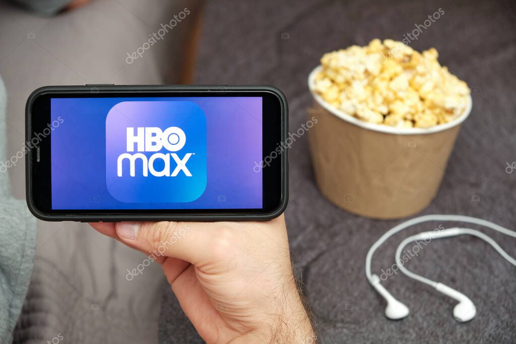 Close up mans hand holding a mobile phone with HBO Max logo with Apple earphones and popcorn box next to him, August 2020, San Francisco, USA.