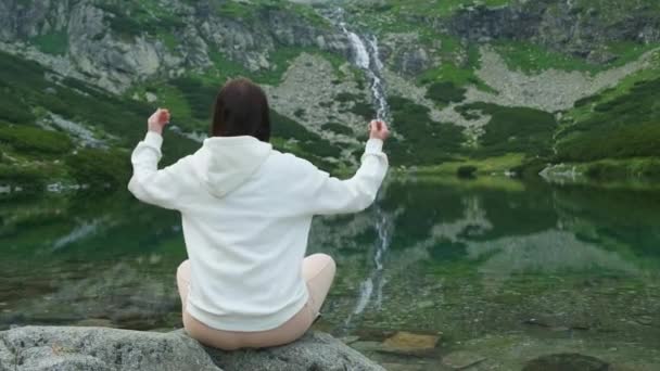 Mujer joven meditando y levantando las manos bajo la cabeza con una vista increíble sobre el lago y la cascada. — Vídeo de stock